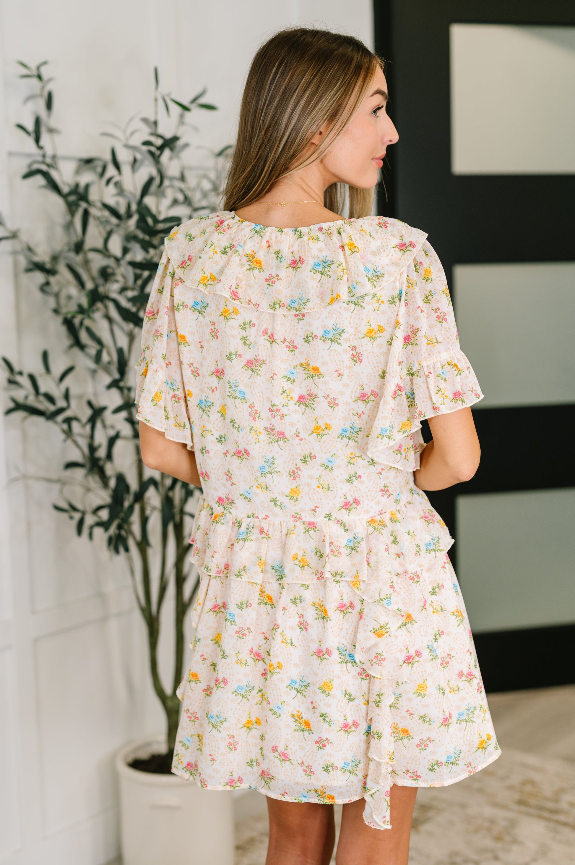 Woman wearing a floral dress standing in a room with a plant and wall art.