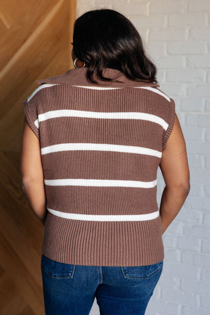 Person wearing a brown sleeveless sweater with white stripes against a white brick wall.
