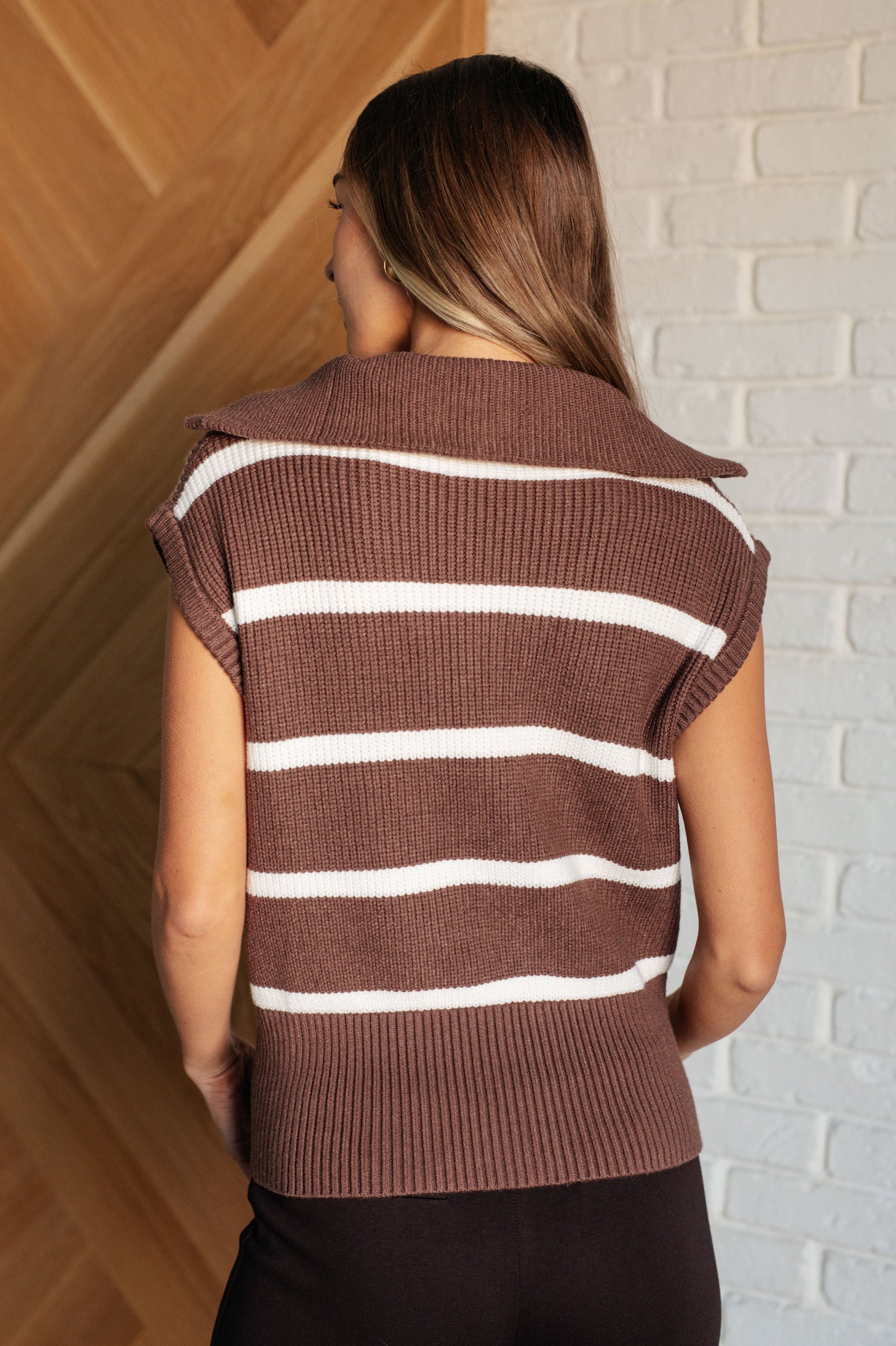 Woman wearing a brown and white striped sleeveless sweater against a brick wall.