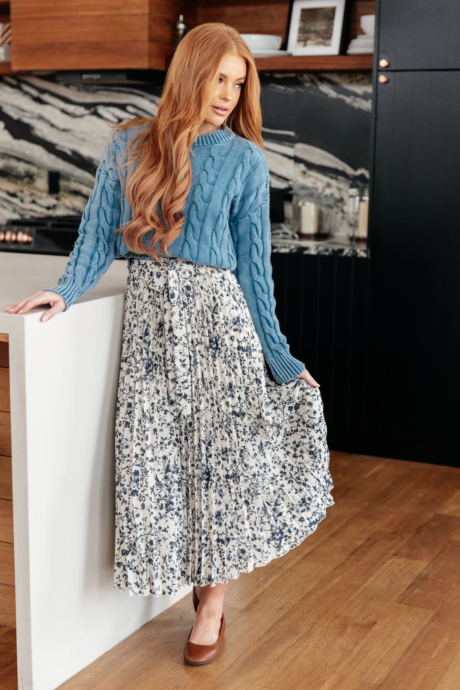 Woman wearing a blue cable knit sweater and patterned skirt in a modern interior setting.