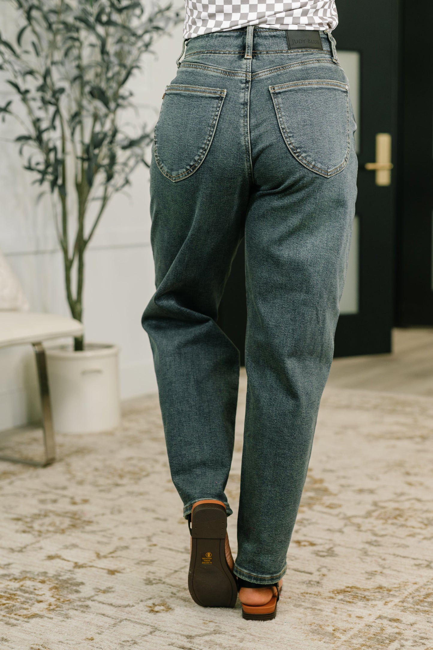 Person wearing blue jeans and brown shoes in an indoor setting with a plant and door in the background.