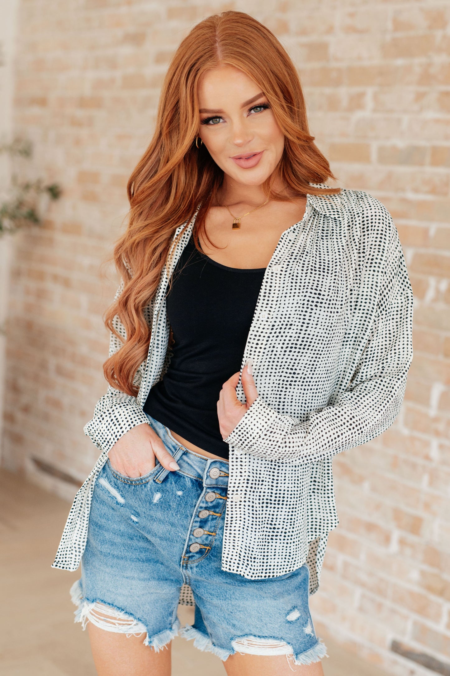 Woman wearing a patterned cardigan, black top, and denim shorts against a brick wall.
