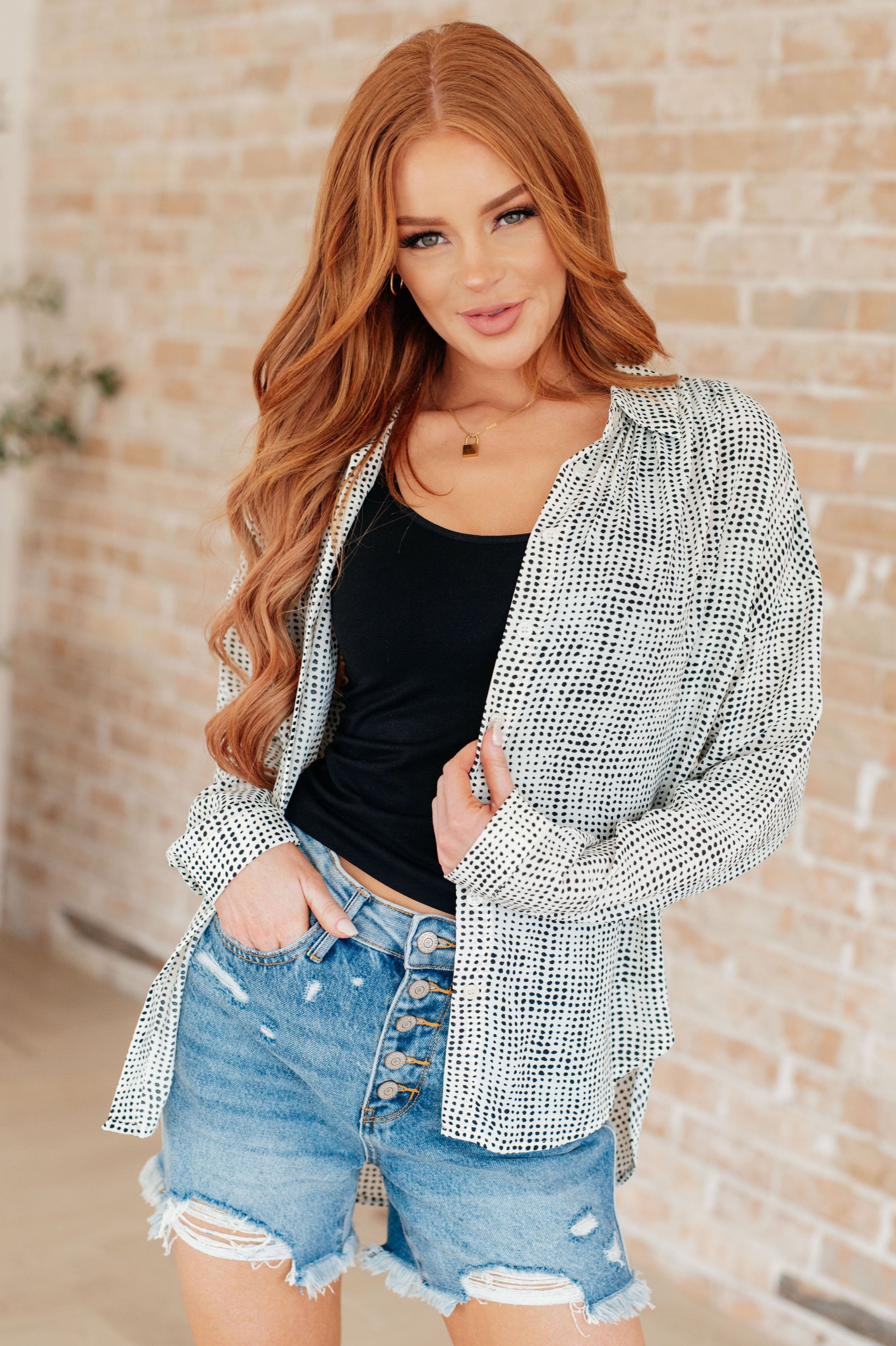 Woman wearing a patterned cardigan, black top, and denim shorts against a brick wall.