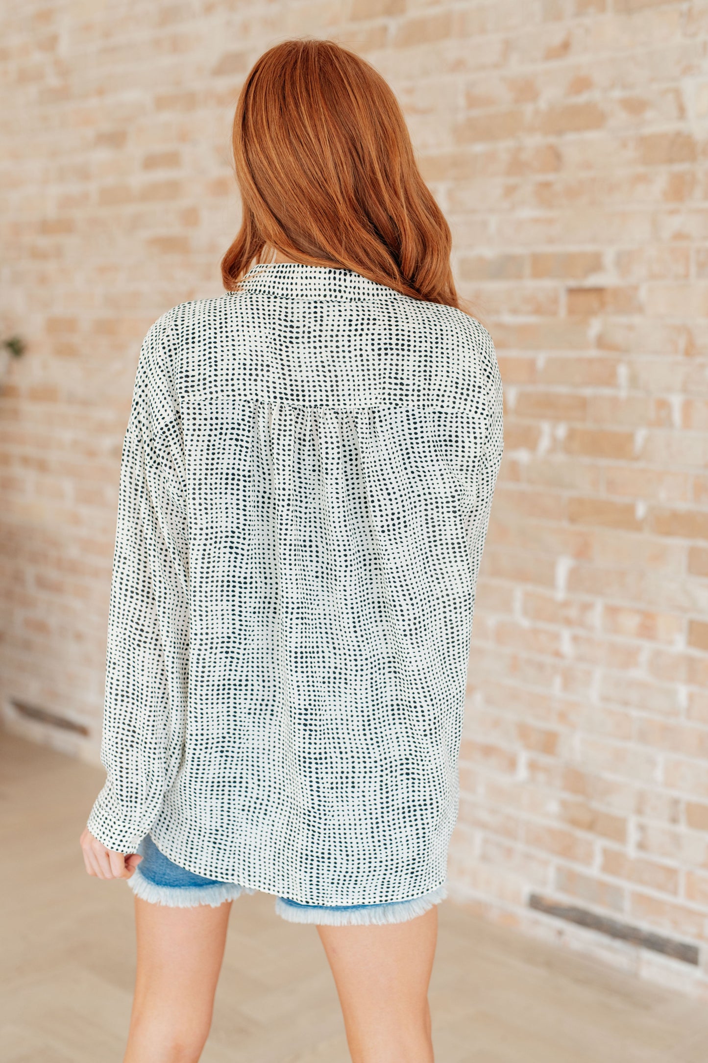 Woman wearing a patterned shirt and denim shorts against a brick wall.