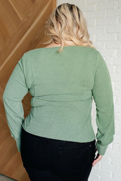 Person wearing a green ribbed sweater against a white brick wall.