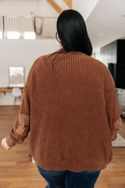 Person wearing a brown ribbed sweater in a room with wooden flooring and a mirror.