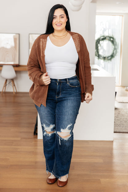 Woman wearing a brown cardigan, white tank top, and blue jeans indoors.