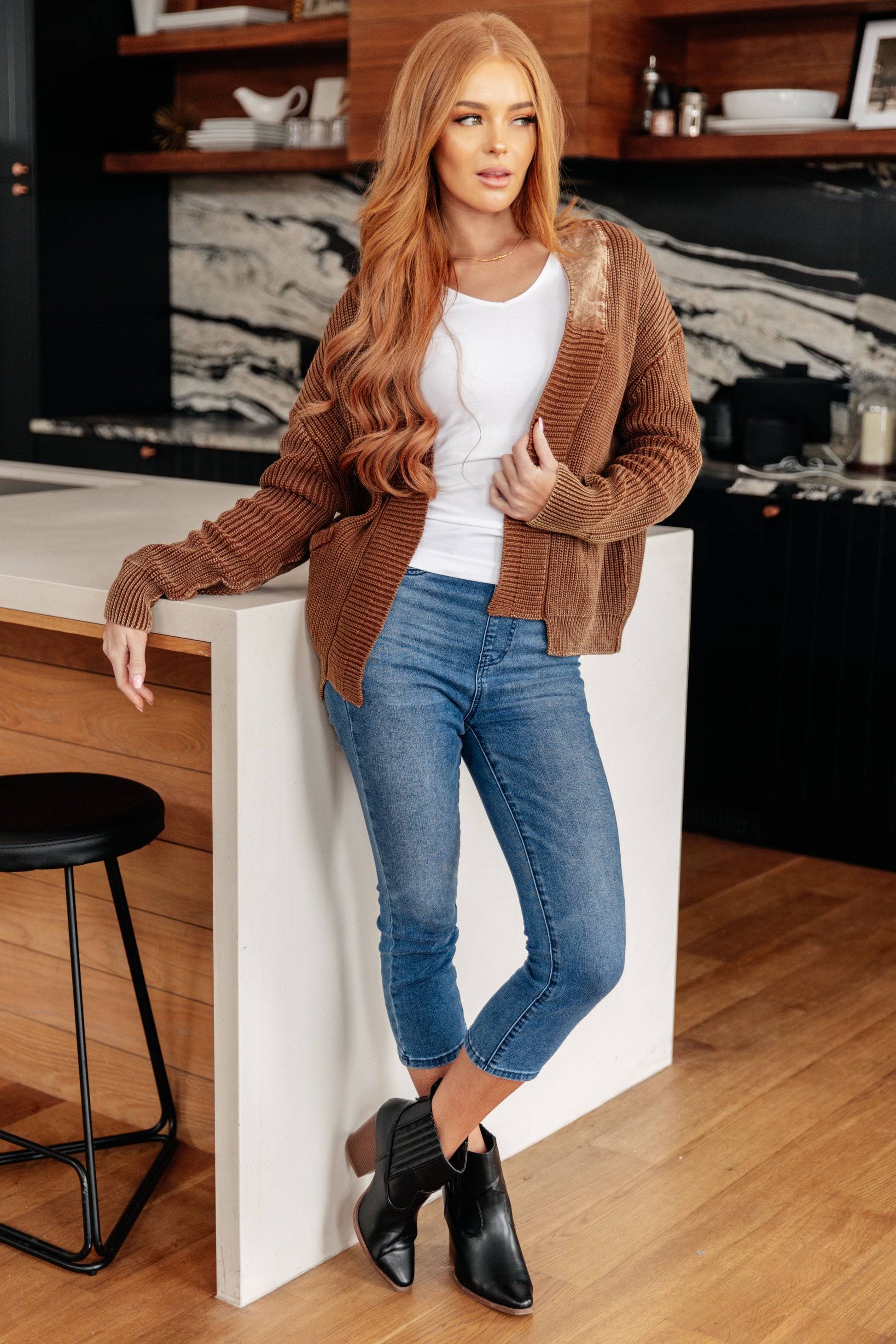 Woman in a brown cardigan, white shirt, blue jeans, and black boots standing in a kitchen.