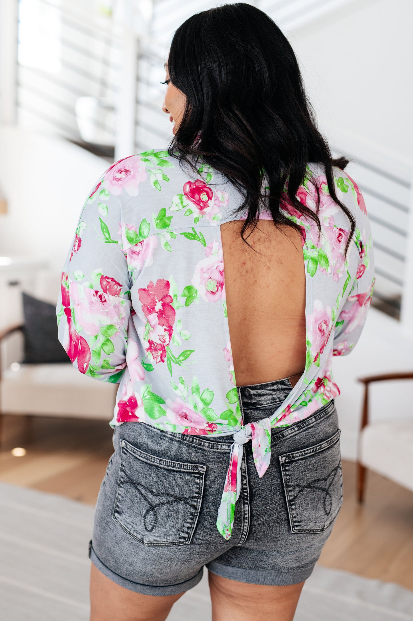 Woman wearing a floral blouse with open back and denim shorts in an indoor setting.