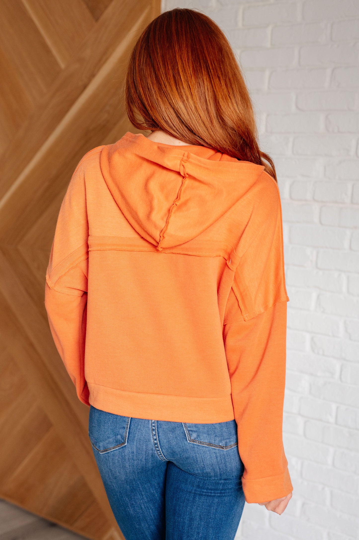 Person wearing an orange hoodie and blue jeans against a white brick wall.