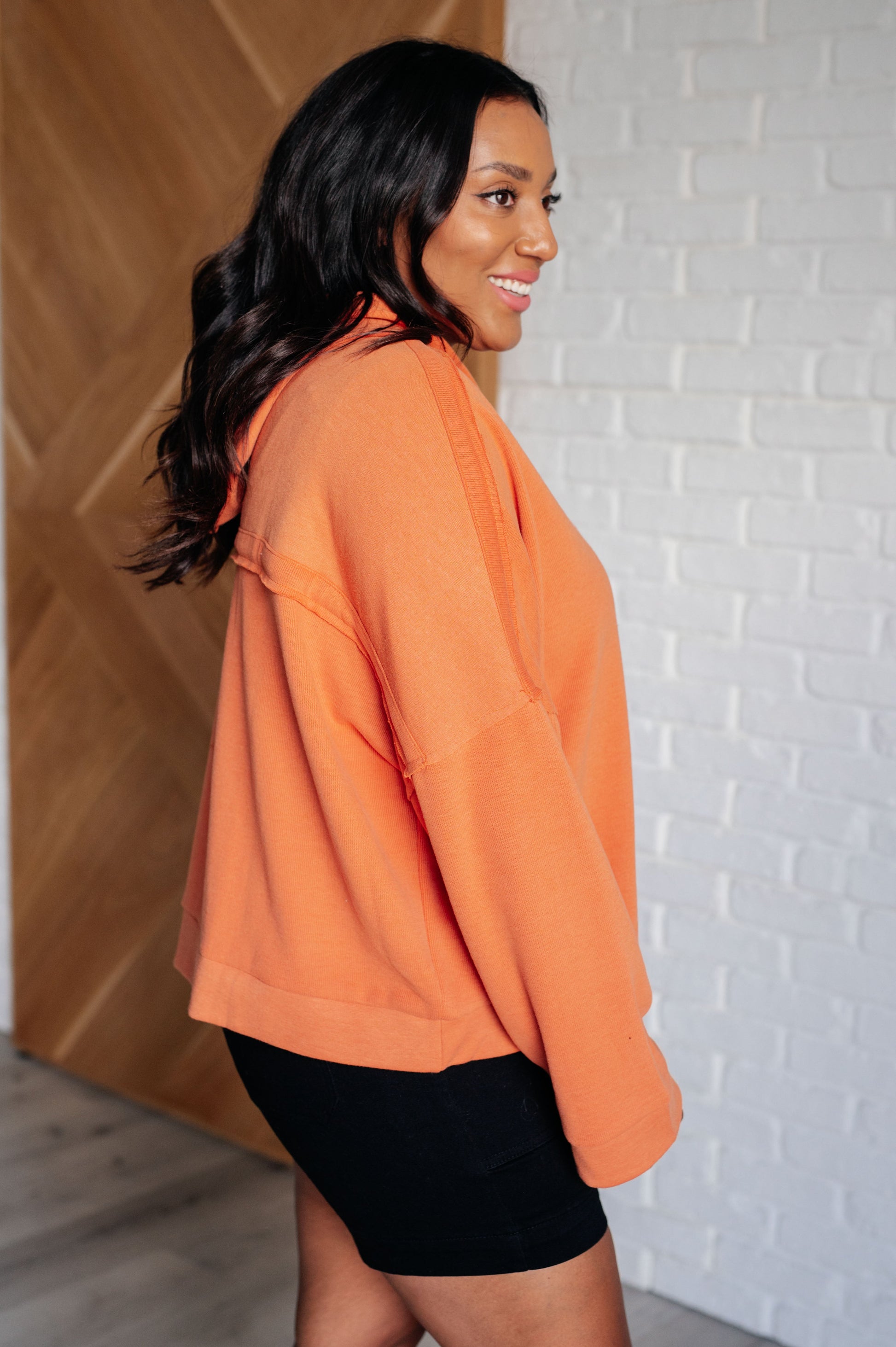 Woman wearing an orange sweatshirt and black shorts against a white brick wall.