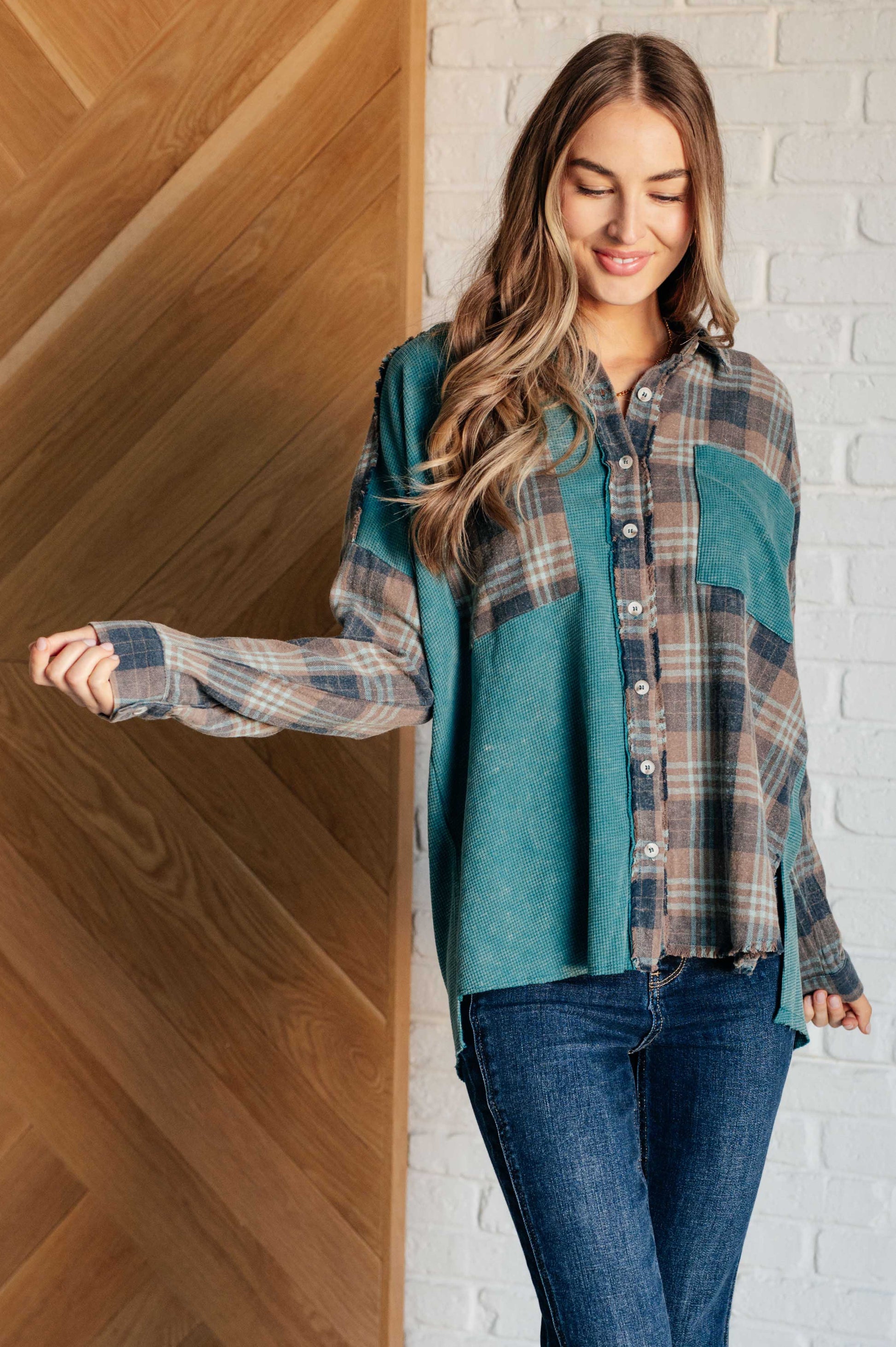 Woman wearing a plaid shirt over a teal base with jeans, standing against a wooden and white brick wall.