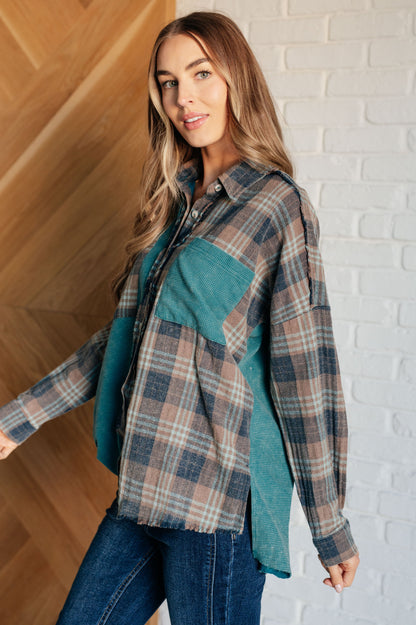 Woman wearing a plaid shirt over a teal top against a white brick wall.