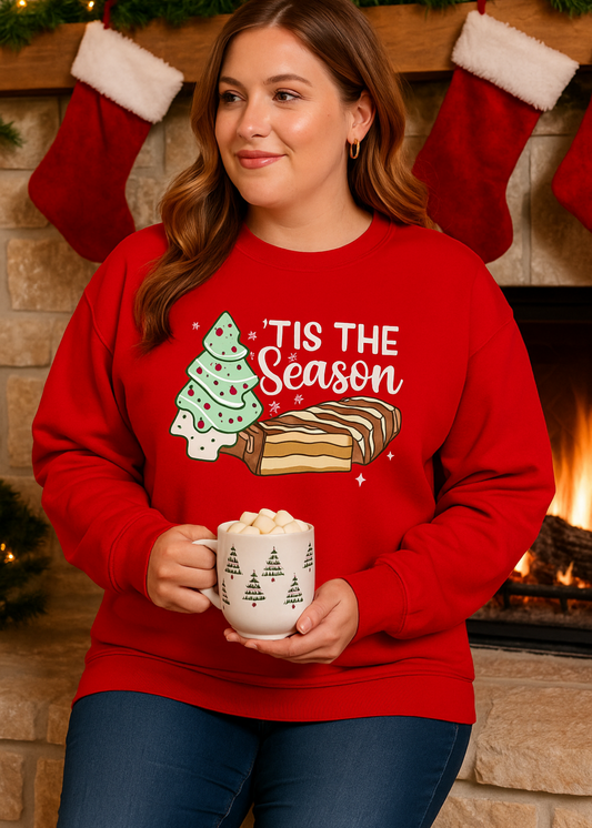 Woman wearing a red 'Tis the Season' sweater holding a mug in front of a fireplace with stockings.