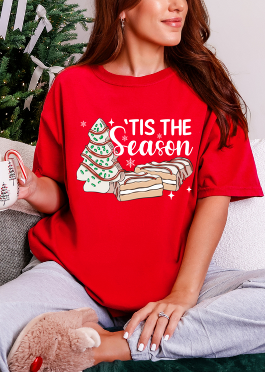 Person wearing a red 'Tis the Season t-shirt with a Christmas tree and cookies design, holding a mug.
