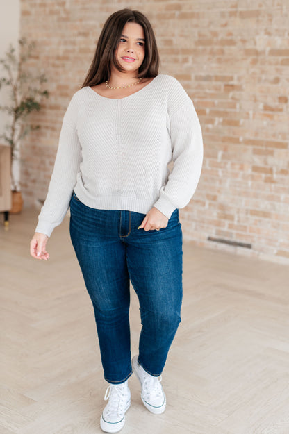 Woman wearing a light gray sweater, blue jeans, and white sneakers in front of a brick wall.