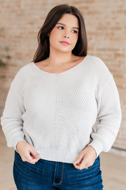 Woman wearing a light gray sweater and blue jeans against a beige wall.