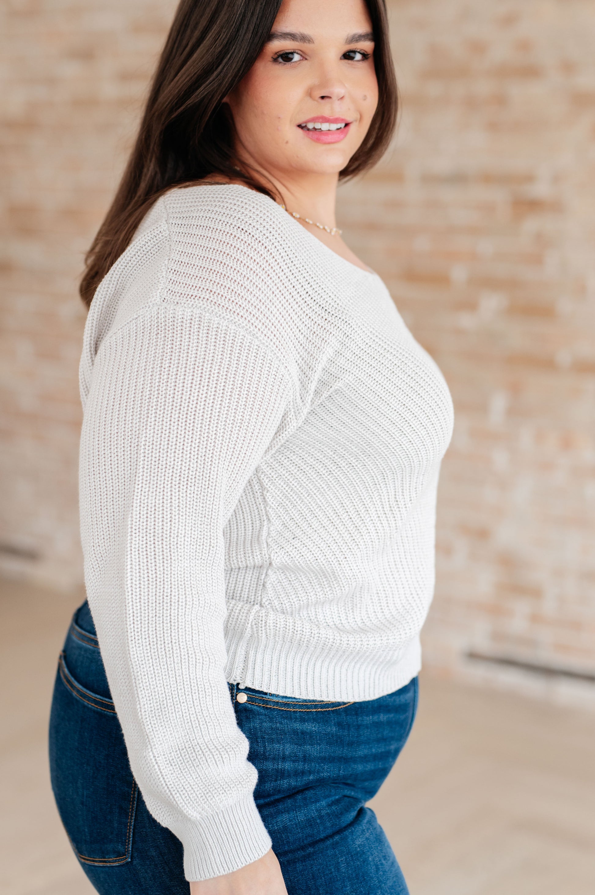 Woman wearing a white sweater and blue jeans against a brick wall.