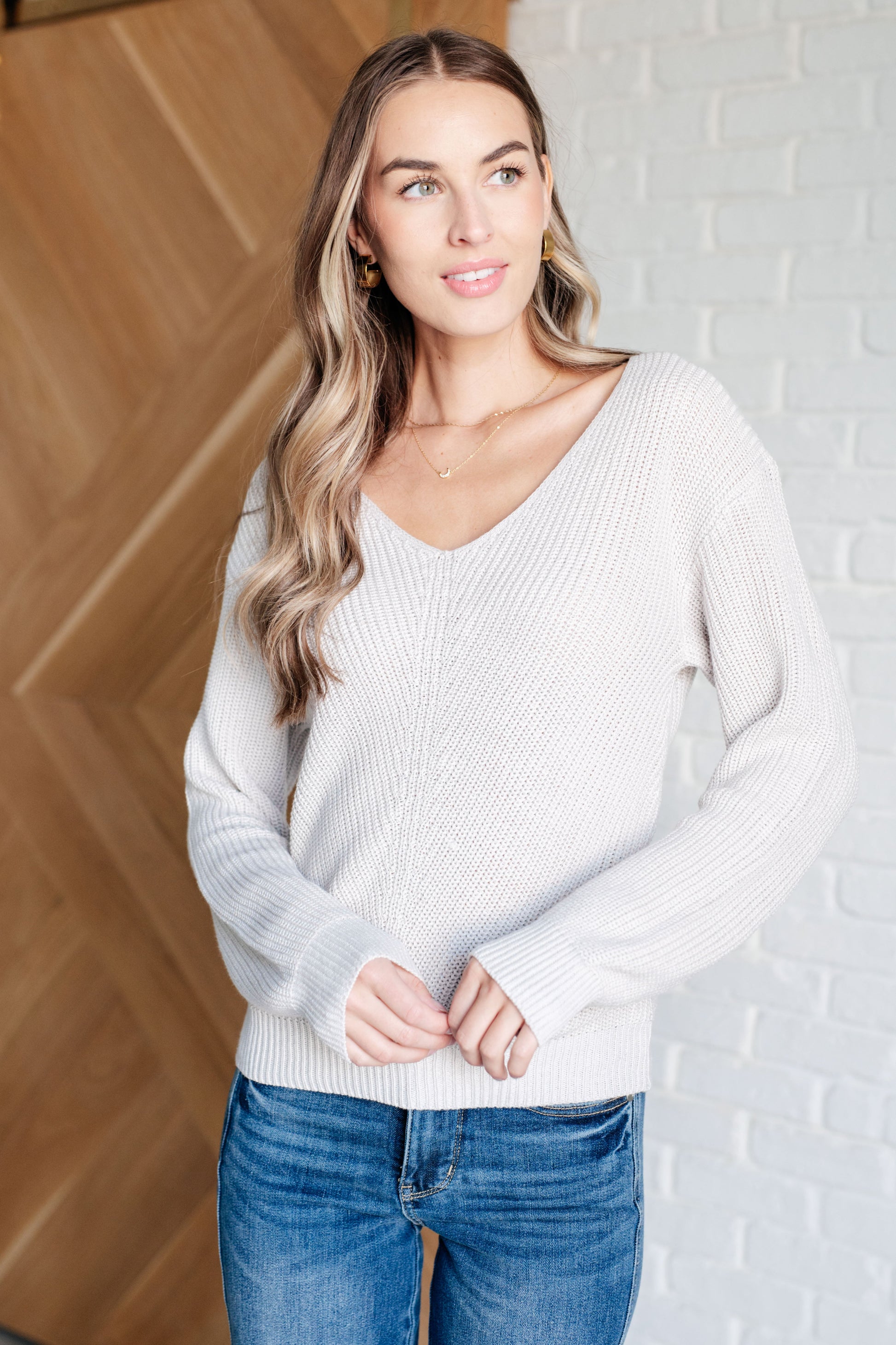 Woman wearing a light gray sweater and blue jeans against a wooden and white brick wall.