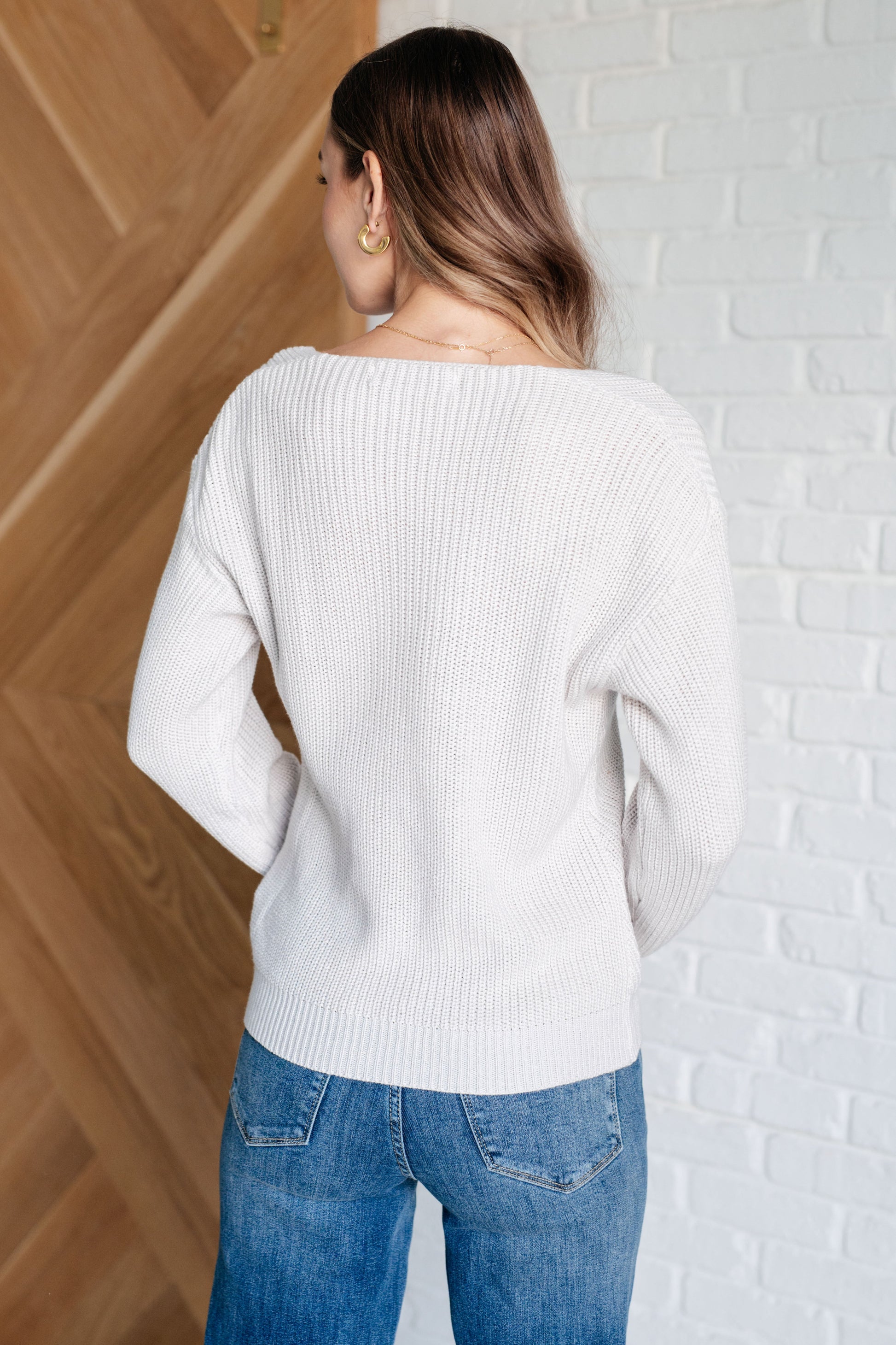 Person wearing a white sweater and blue jeans against a wooden and white brick wall.