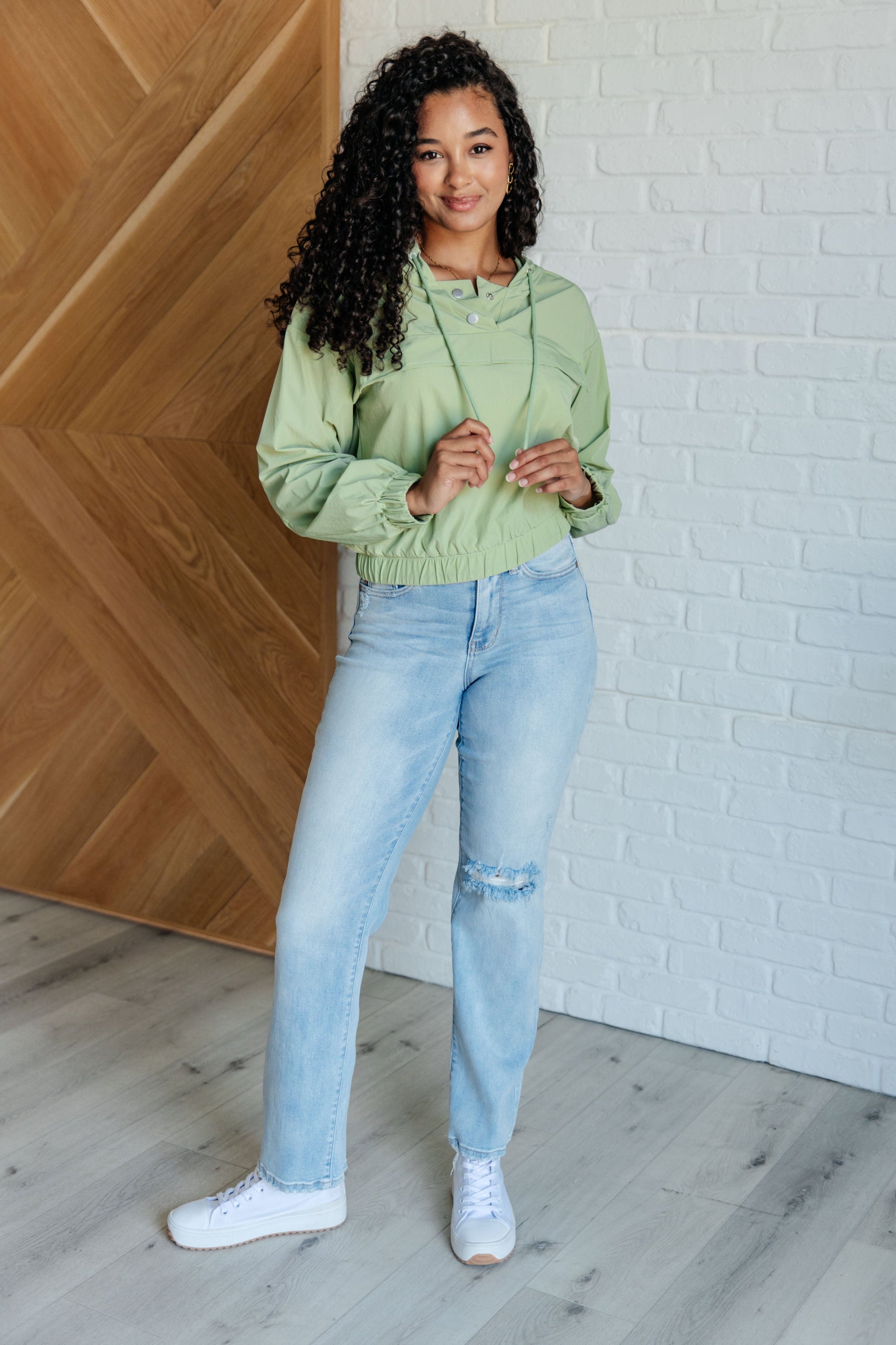 Person wearing a light green hoodie and blue jeans standing against a wooden and white brick wall.