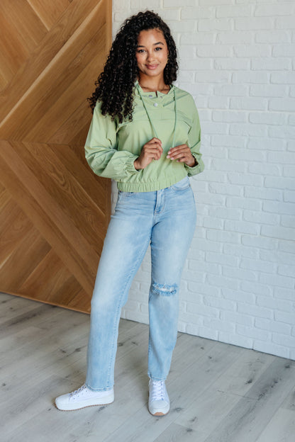 Person wearing a light green hoodie and blue jeans standing against a wooden and white brick wall.