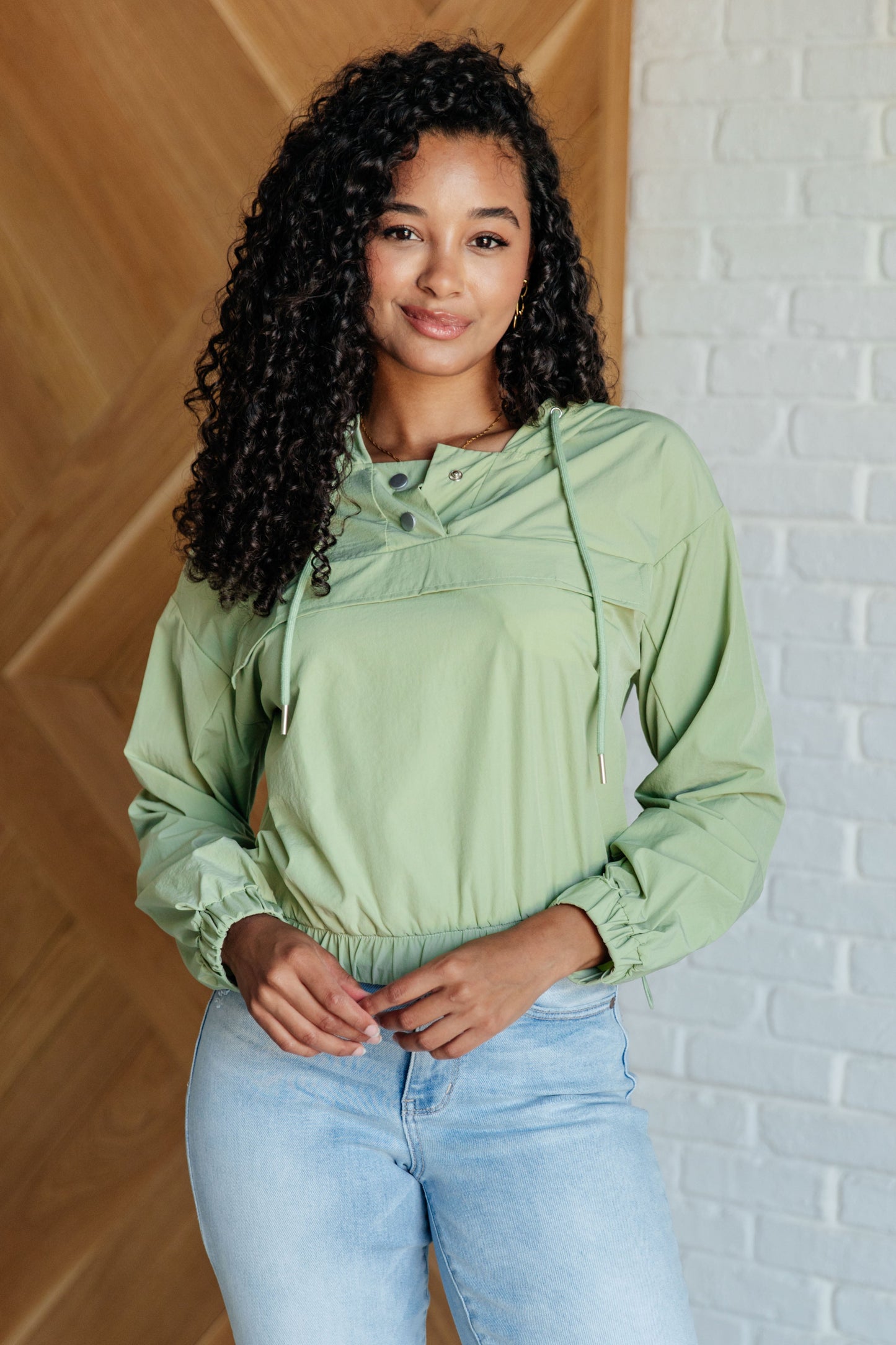 Woman wearing a green top and blue jeans standing against a white brick wall.