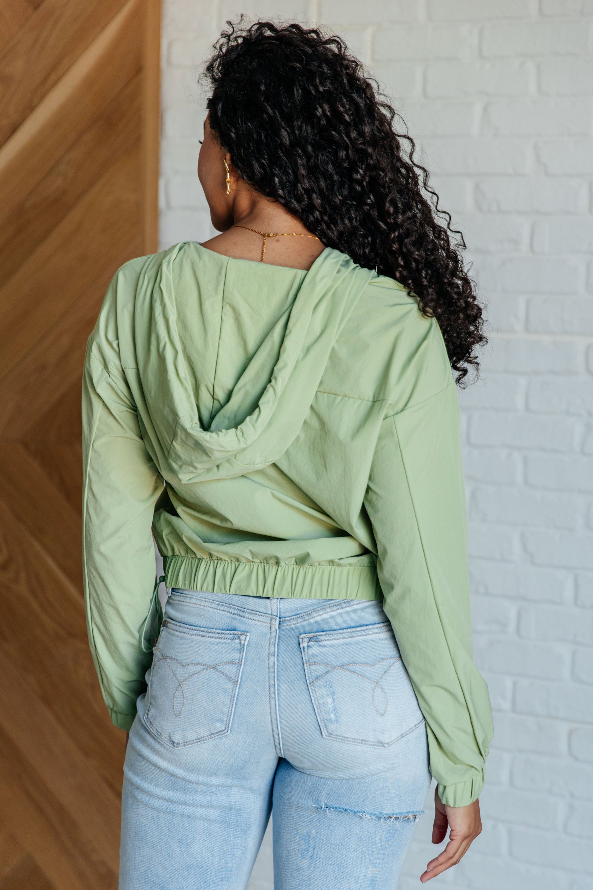 Person wearing a light green jacket and light blue jeans against a white brick wall.