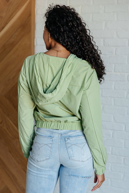 Person wearing a light green jacket and light blue jeans against a white brick wall.