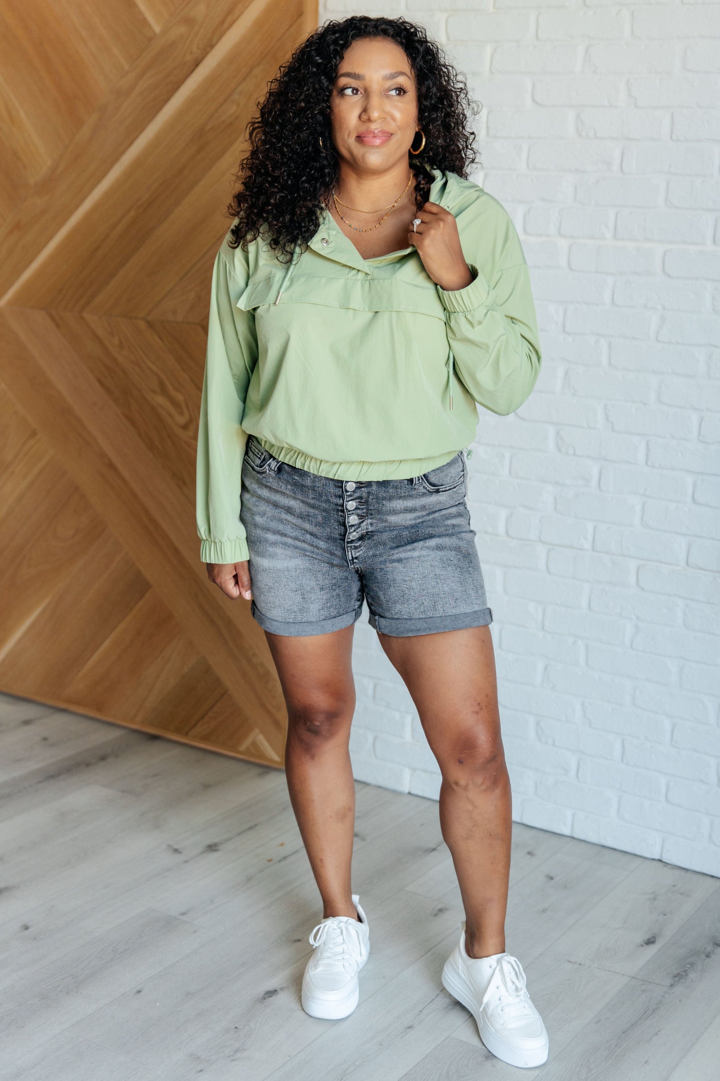 Woman wearing a light green hoodie and denim shorts standing against a white brick wall.