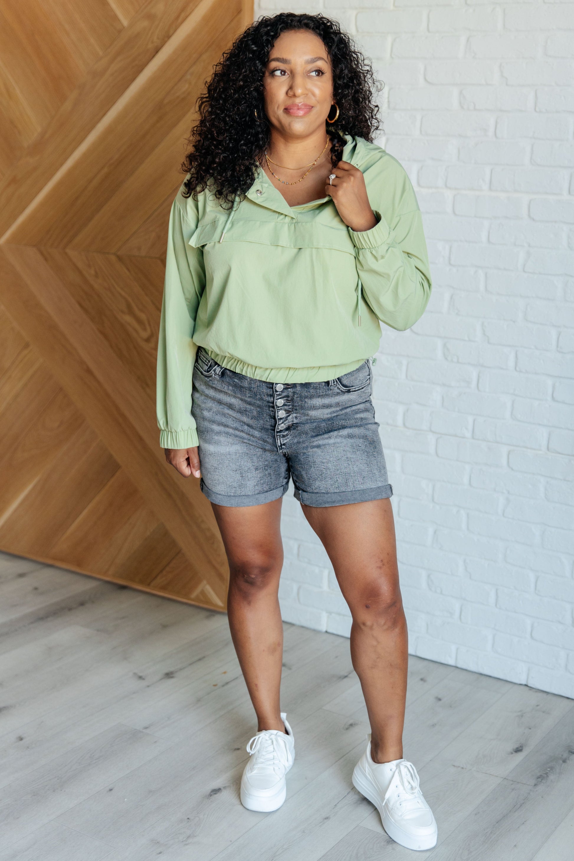 Woman wearing a light green hoodie and denim shorts standing against a white brick wall.