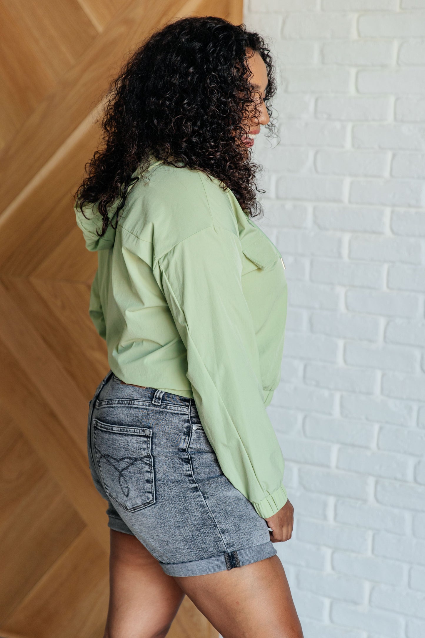 Person wearing a light green hoodie and denim shorts against a white brick wall.