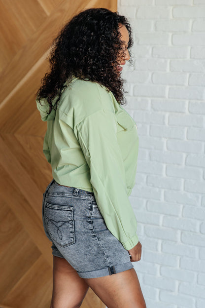 Person wearing a light green hoodie and denim shorts against a white brick wall.