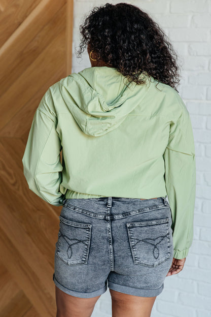 Person wearing a light green jacket and denim shorts standing against a white brick wall.