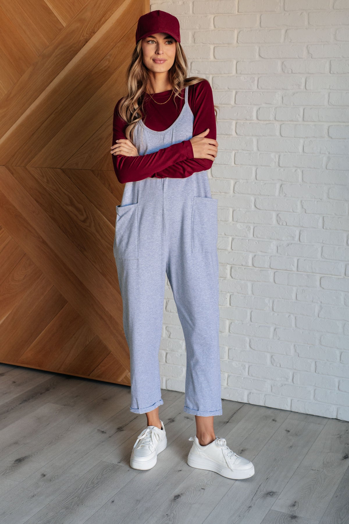 Woman wearing a red top and light blue overalls standing against a white brick wall.
