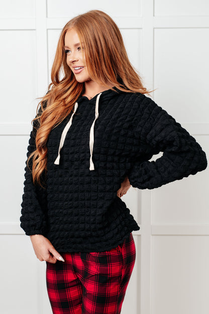 Woman wearing a black textured hoodie and red plaid pants against a white wall.