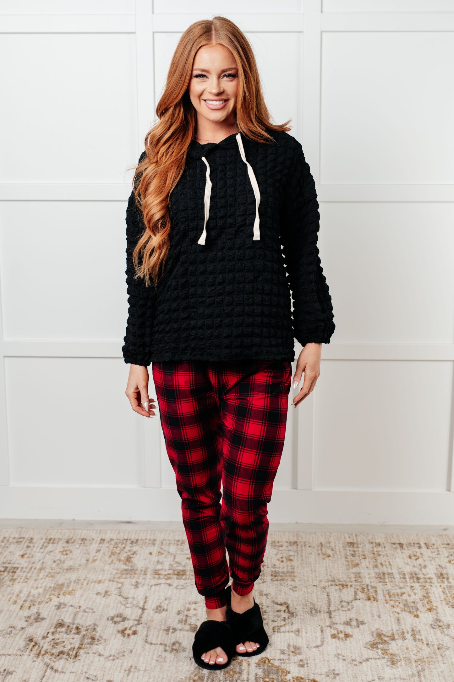 Woman wearing a black hoodie and red plaid pants standing against a white wall.
