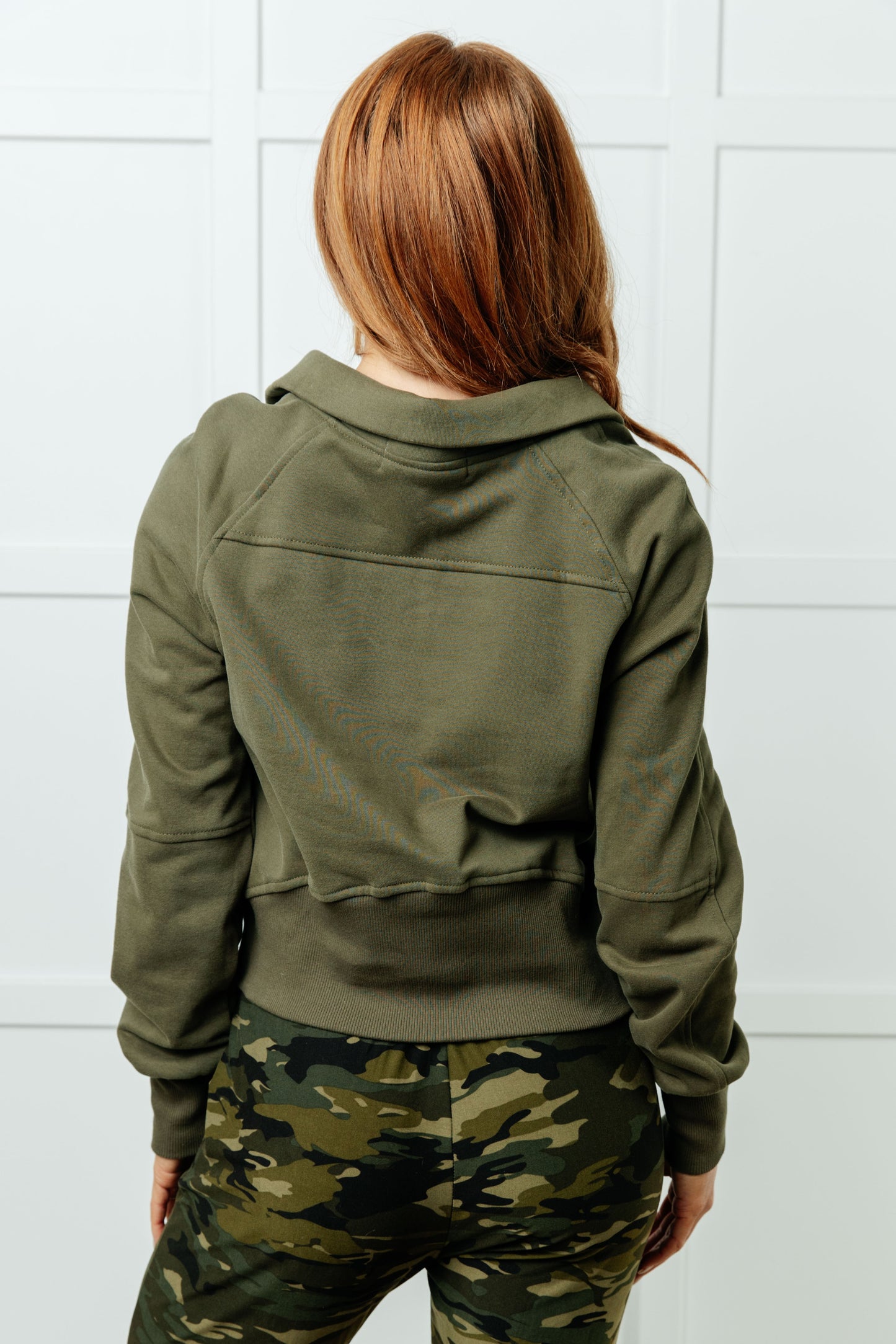 Person wearing a green jacket and camouflage pants against a white tiled wall.