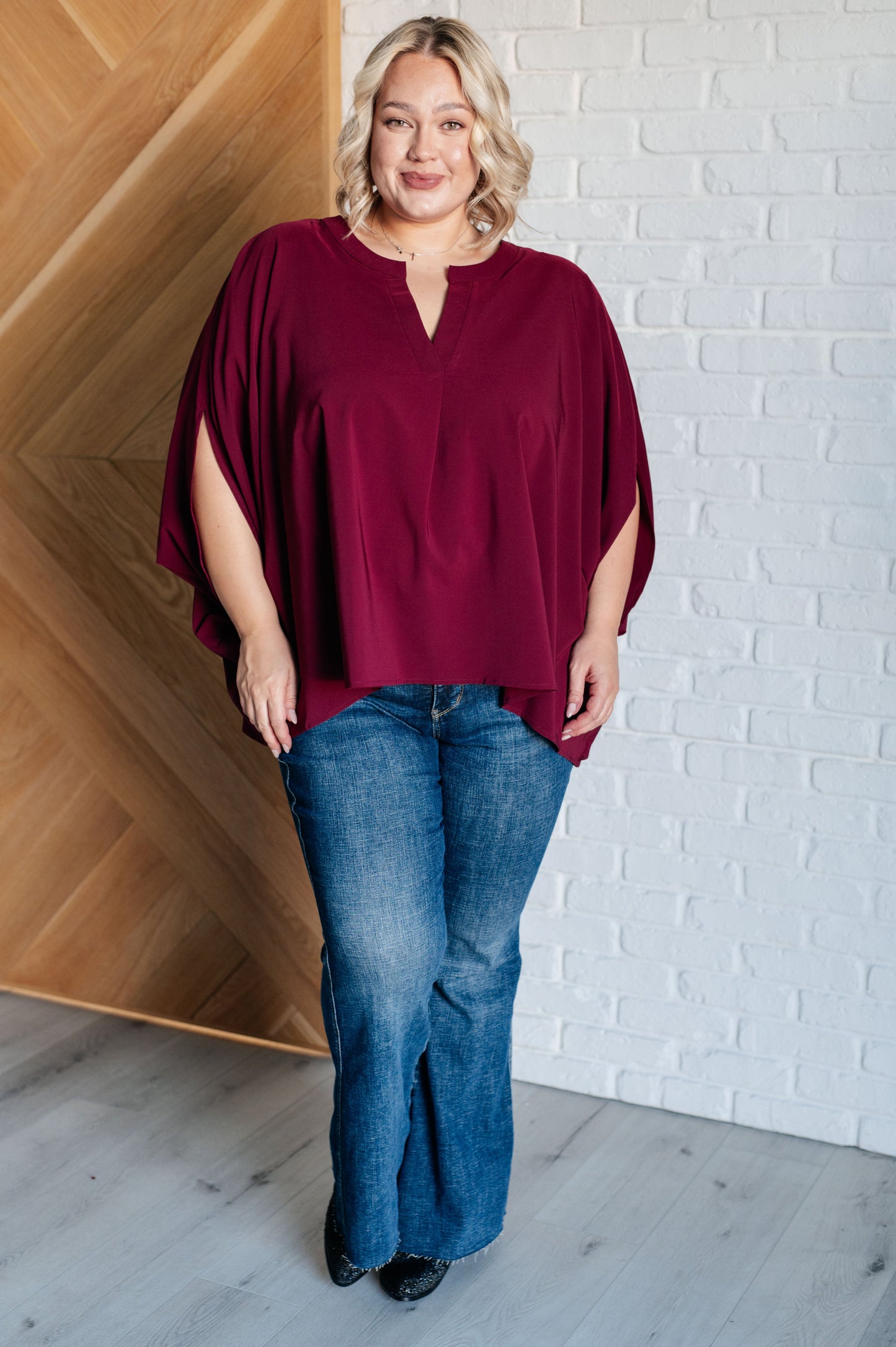 Woman wearing a burgundy top and blue jeans standing against a white brick wall.
