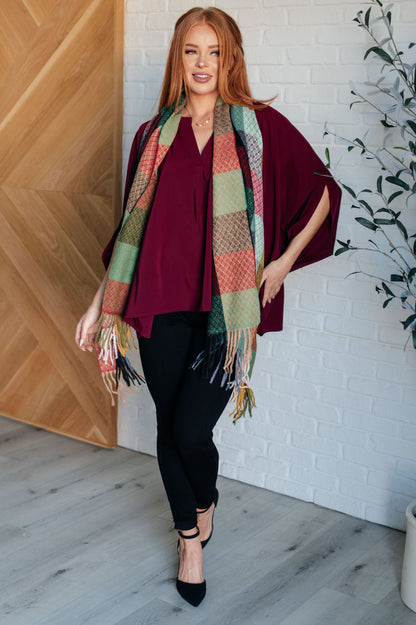 Woman wearing a burgundy top, black pants, and a patterned scarf in a room with wooden and white brick walls.
