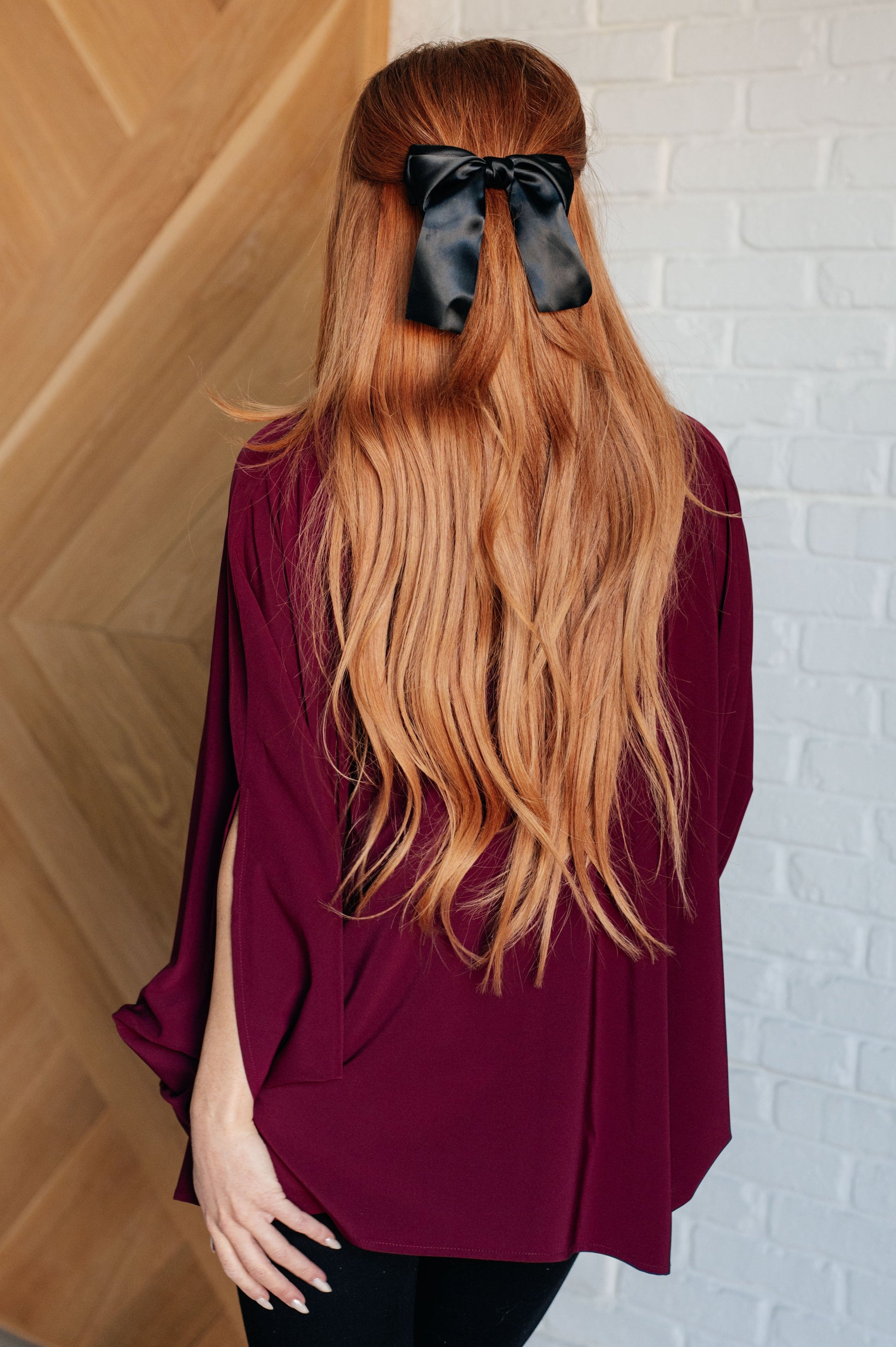 Person wearing a burgundy top with long blonde hair tied back with a black bow, standing against a white brick wall.