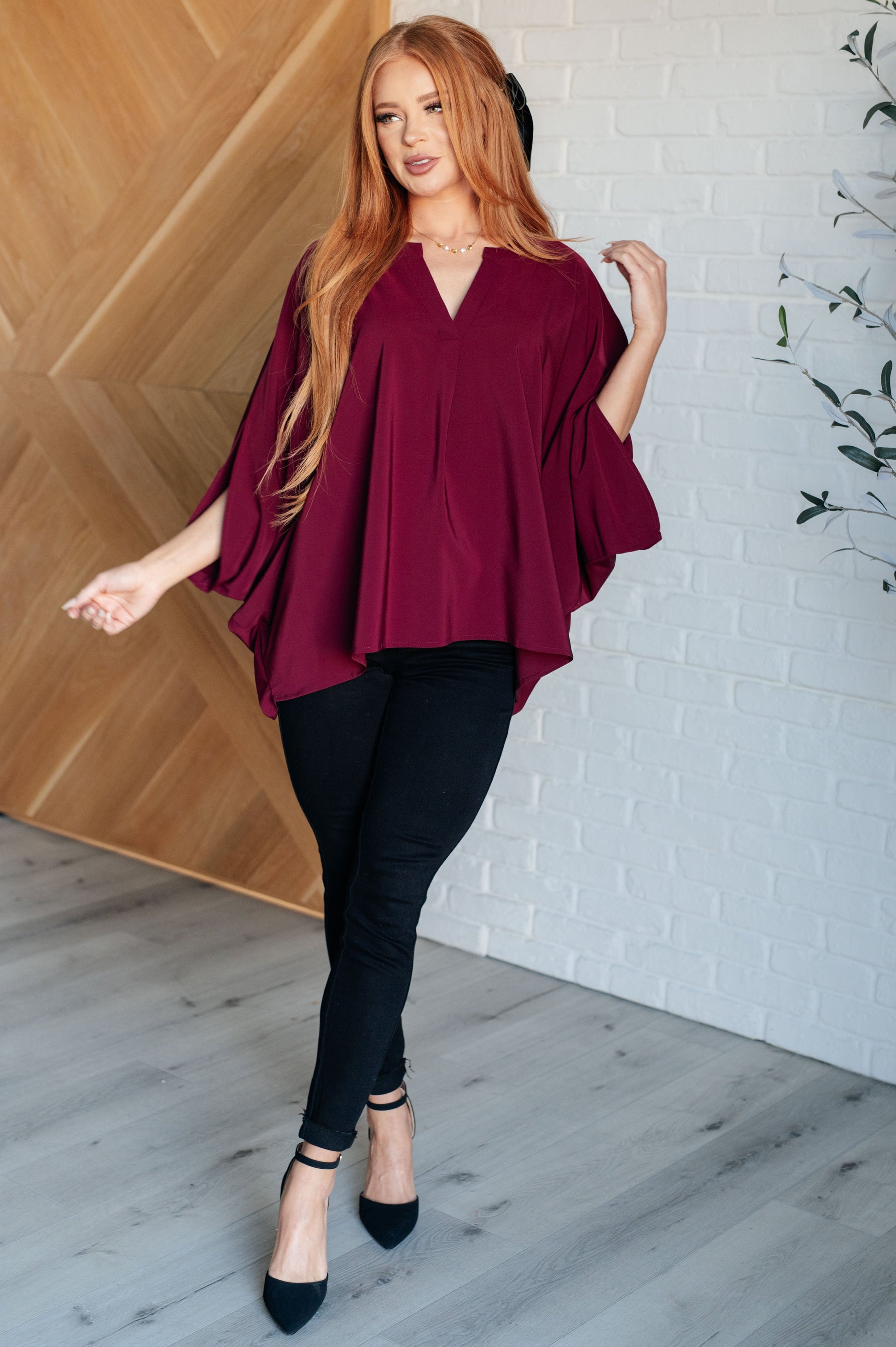 Woman wearing a burgundy top and black pants standing in a room with wooden and white brick walls.