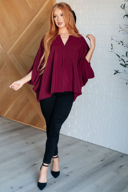 Woman wearing a burgundy top and black pants standing in a room with wooden and white brick walls.