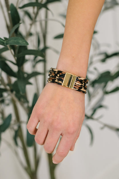 Hand wearing a leopard print and gold bracelet with a blurred green plant background