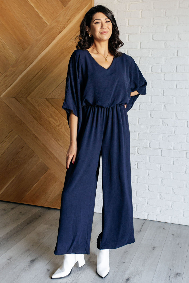 Woman wearing a navy blue jumpsuit standing against a white brick wall.