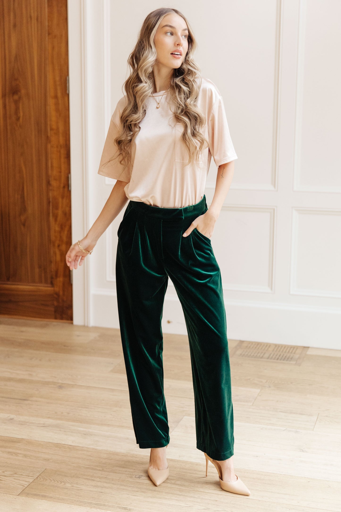 Woman wearing a beige top and green velvet pants in a room with wooden floor and white walls.