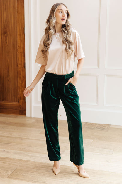Woman wearing a beige top and green velvet pants in a room with wooden floor and white walls.