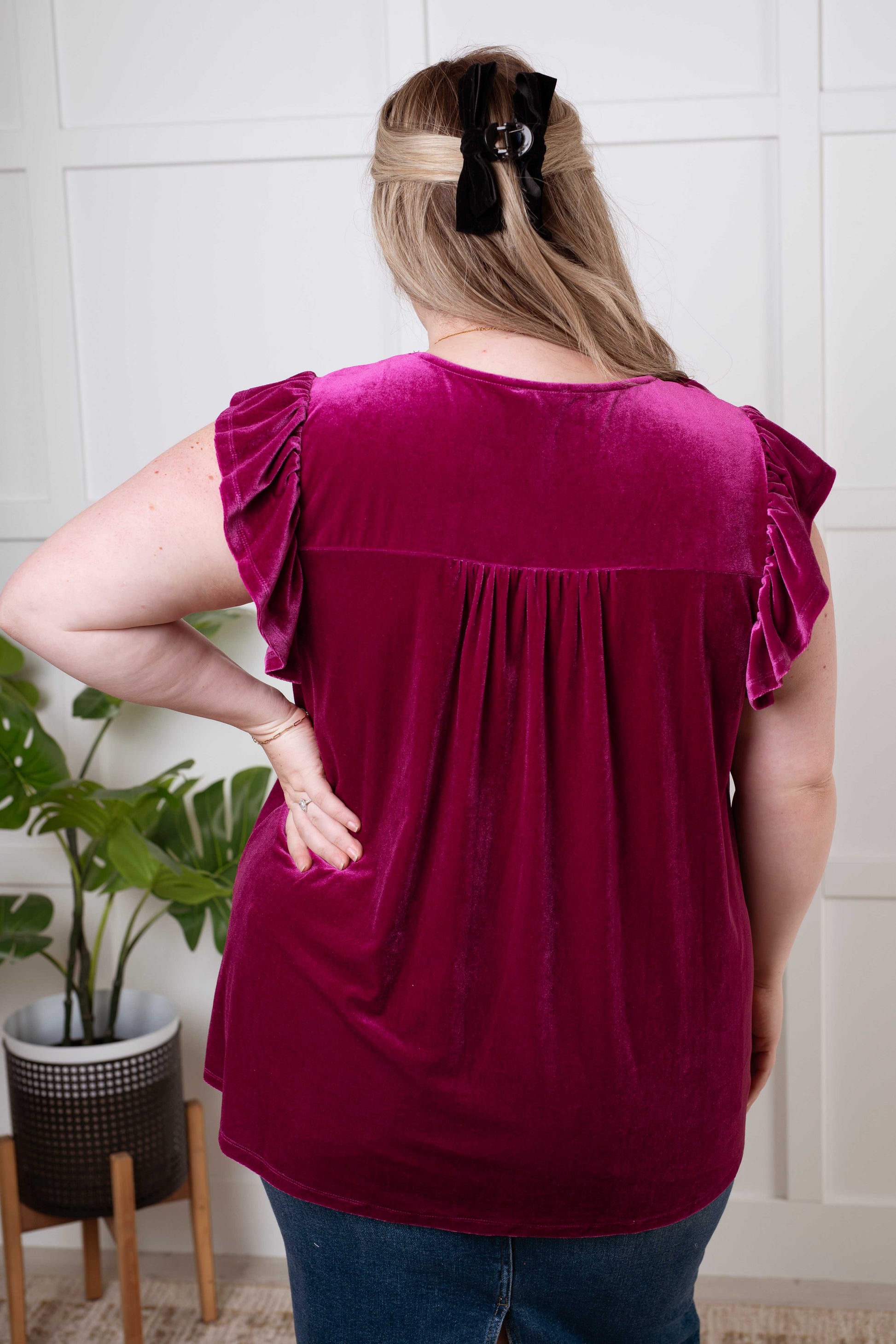 Person wearing a burgundy top with ruffled sleeves in a room with a plant and tiled wall.