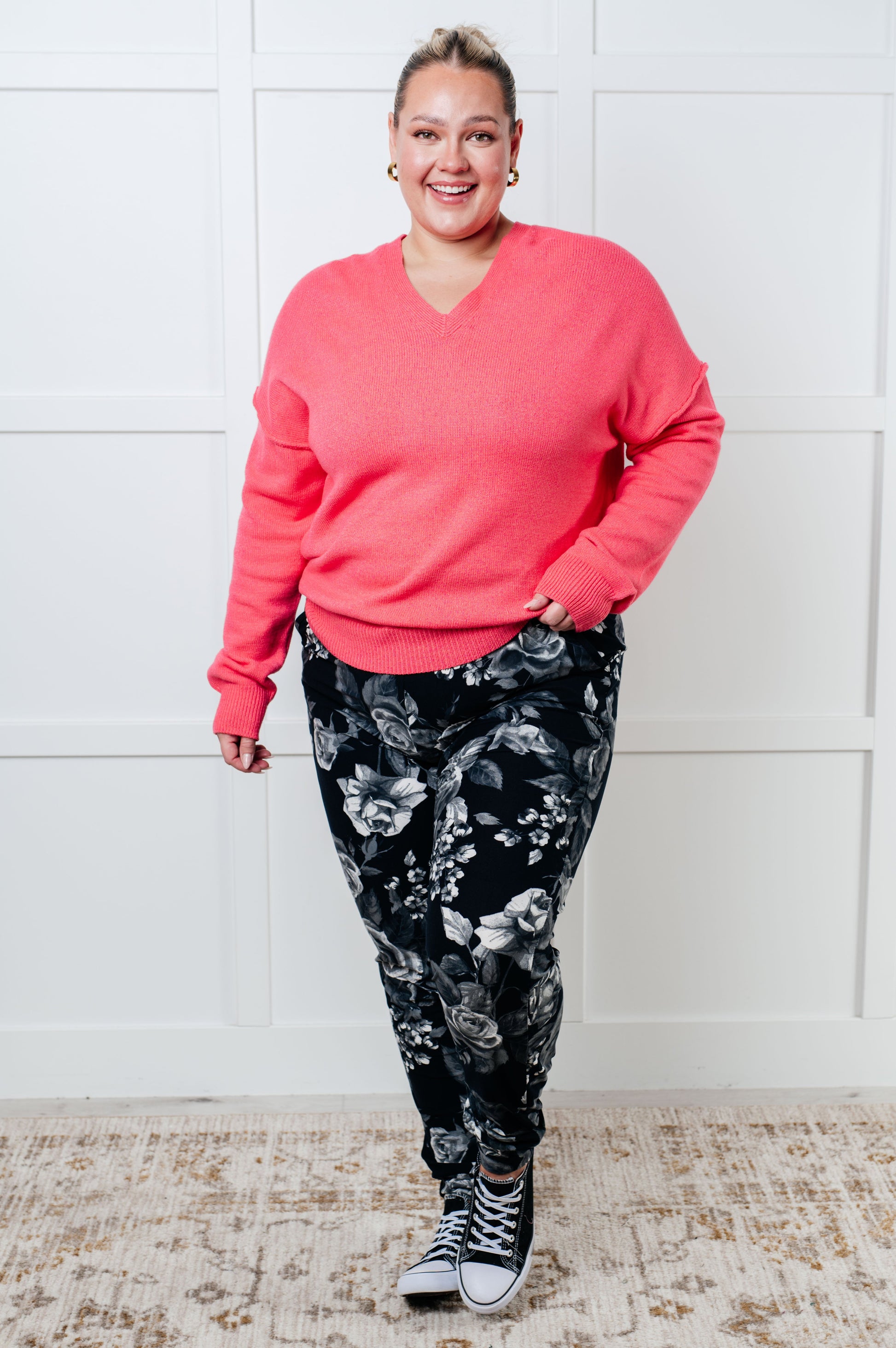 Person wearing a pink sweater and black floral pants against a white wall.