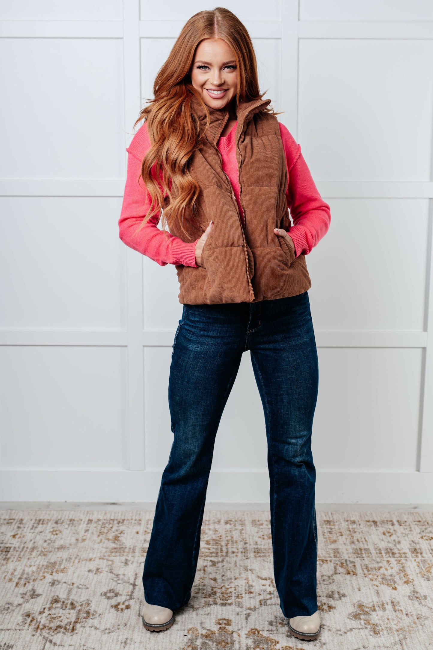 Woman wearing a brown vest over a pink shirt with blue jeans against a white paneled wall.